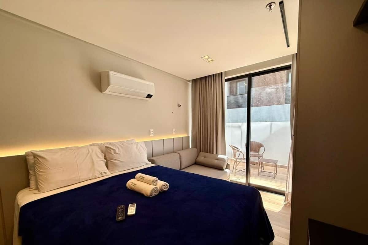 A hotel room with a bed, a bedside table, a remote control, a window, and a view of the outside.