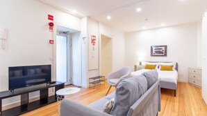 Ett modernt hotellrum med en platt-TV, en soffa, en säng med gula kuddar och ett litet sidobord.