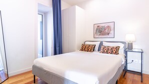 Ein Hotelzimmer mit einem Bett, einem Nachttisch mit Lampe, einem Gemälde an der Wand und einer Tür zu einem anderen Raum.