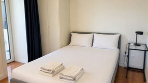 Ein ordentlich bezogenes Bett mit zwei Kissen, ein kleiner Nachttisch mit Lampe und ein Fenster mit Vorhängen.