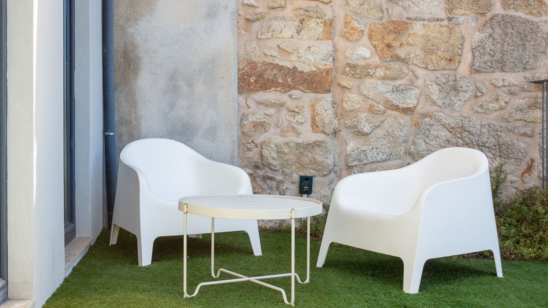 A white outdoor seating area with a table and two chairs, set against a stone wall and a grassy surface.