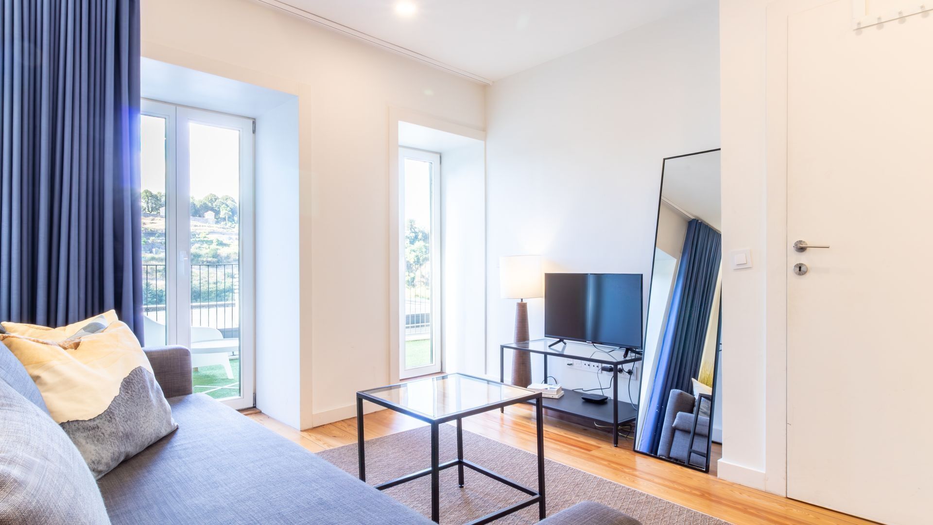 Un salon moderne avec un canapé, une table basse en verre, un téléviseur à écran plat et un balcon agrémenté de rideaux.