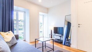 A modern living room with a sofa, a glass coffee table, a flat-screen TV, and a balcony with curtains. - E: Sweeping Panoramic Terrace | Douro Views (Vila Nova de Gaia)