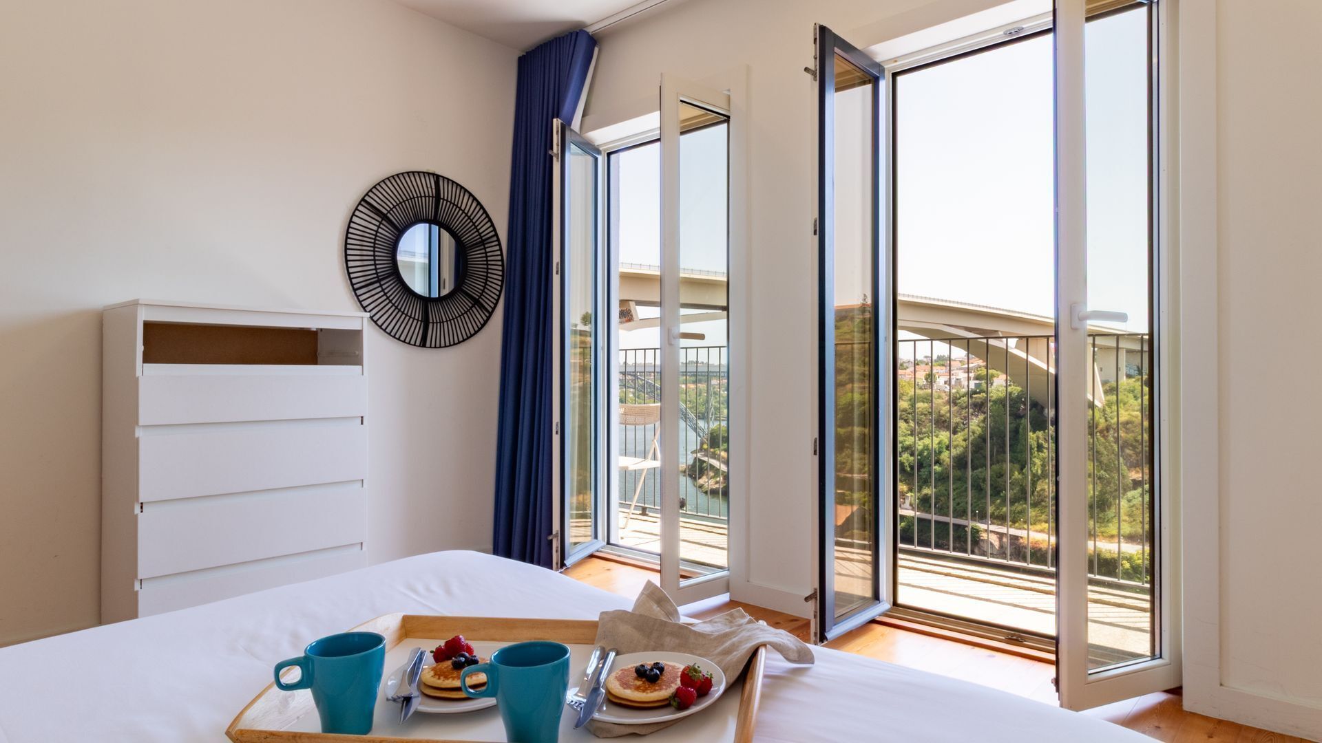 A bedroom with a bed, a breakfast tray with pancakes and berries, a large window with a view of a bridge and greenery, and a wall-mounted mirror.