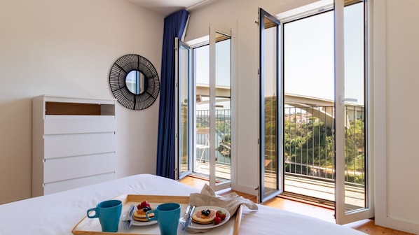 A bedroom with a bed, a breakfast tray with pancakes and berries, a large window with a view of a bridge and greenery, and a wall-mounted mirror. - F: Ponte Luis Balcony | Riverside Flat (Vila Nova de Gaia)