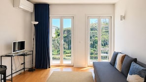 A modern living room with a flat-screen TV, a sofa, a desk with a computer, and a large window with a view of greenery. - F: Ponte Luis Balcony | Riverside Flat (Vila Nova de Gaia)