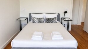 A neatly made bed with white linens and two heart-patterned pillows, a bedside table with a lamp, and a power outlet on the wall. - F: Ponte Luis Balcony | Riverside Flat (Vila Nova de Gaia)