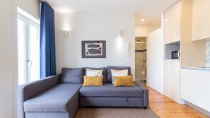 A modern living room with a grey sectional sofa, a small kitchen area, and a visible shower. - G: Iconic Bridge View Balcony | Spacious 1BR Porto (Vila Nova de Gaia)