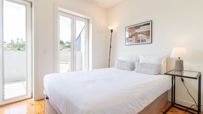 A bedroom with a bed, a nightstand, a lamp, a painting, and a sliding glass door leading to an outdoor area. - G: Iconic Bridge View Balcony | Spacious 1BR Porto (Vila Nova de Gaia)