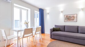 Ett modernt vardagsrum med en grå soffa, vita stolar, ett matbord och ett stort fönster med blå gardiner.