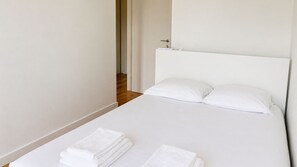 Ein ordentlich bezogenes Bett mit weißen Leinen und Kissen, eine Tür zu einem anderen Raum und ein Holzboden.