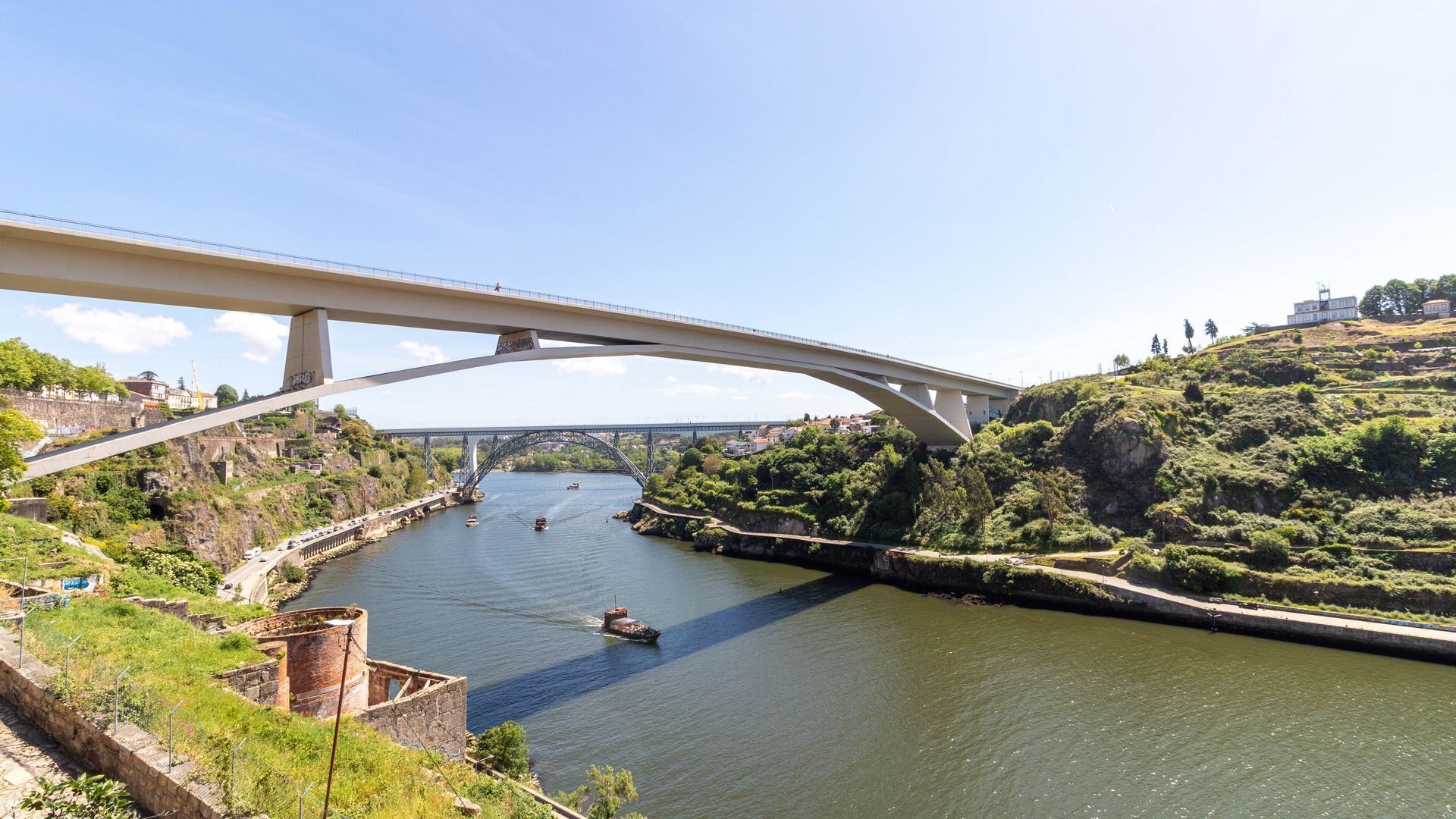 A bridge over a river with boats, greenery, and buildings in the background.