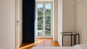 A room with a bed, a small table, and a view of a hillside through a glass door. - J: Iconic Bridge & Douro Views | 1BR Porto (Vila Nova de Gaia)