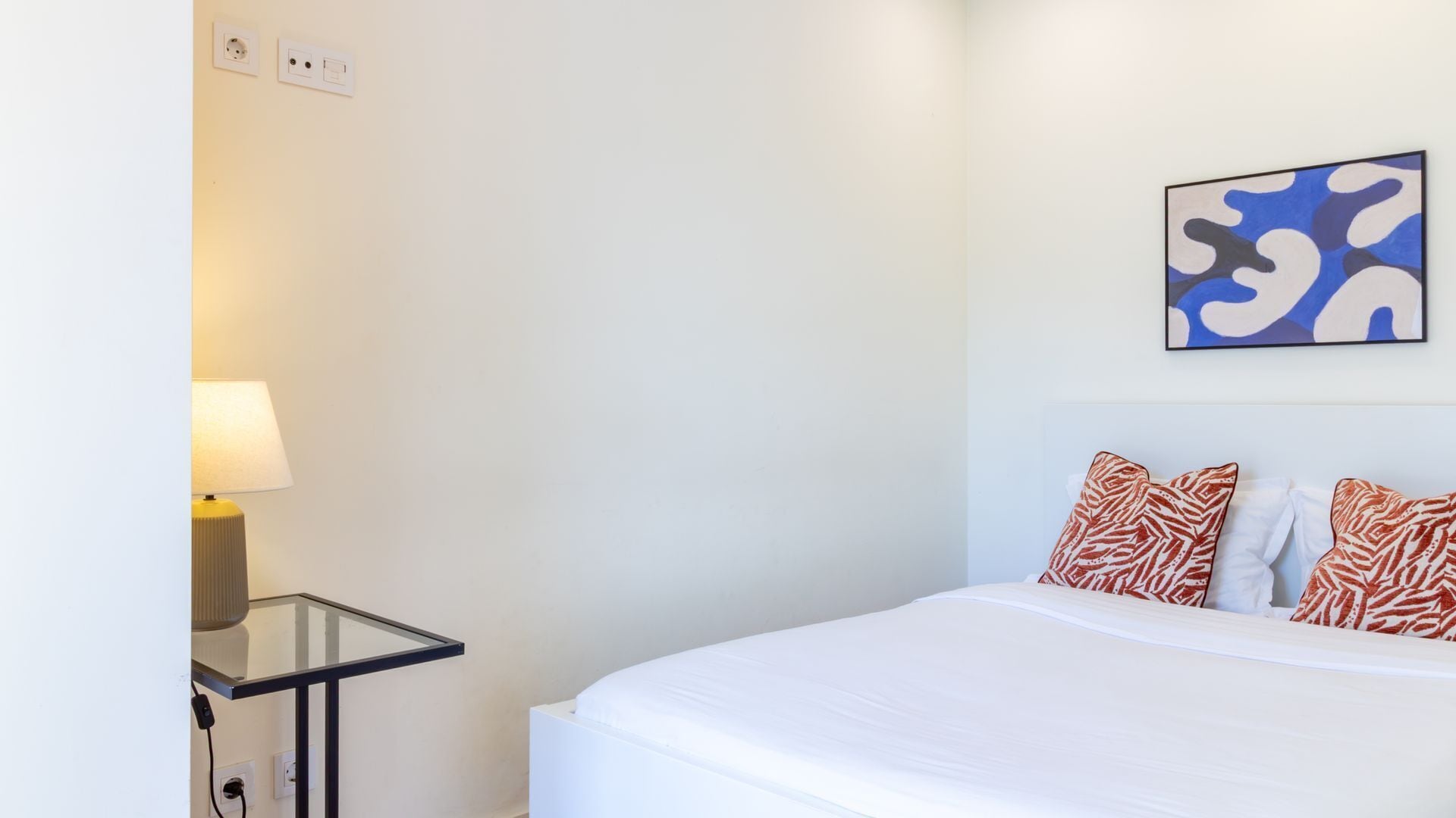 A hotel room with a bed, a bedside table with a lamp, a power outlet, and a wall-mounted abstract painting.