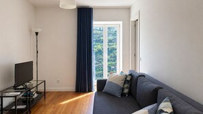 A modern living room with a sofa, a TV on a stand, a tall floor lamp, and a large window with a view of greenery. - J: Iconic Bridge & Douro Views | 1BR Porto (Vila Nova de Gaia)