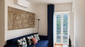 A living room with a stone wall, a blue sofa with patterned cushions, a floor lamp, and a sliding glass door leading to a balcony. - O: Douro & Bridge Views | Boutique Apartments (Vila Nova de Gaia)