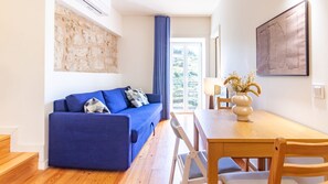 A modern living room with a blue sofa, a wooden dining table, and a view through a large window. - O: Douro & Bridge Views | Boutique Apartments (Vila Nova de Gaia)