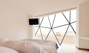 Makuuhuone, jossa on sänky, seinälle asennettu televisio ja suuri ikkuna, jossa on geometrinen kuvio.