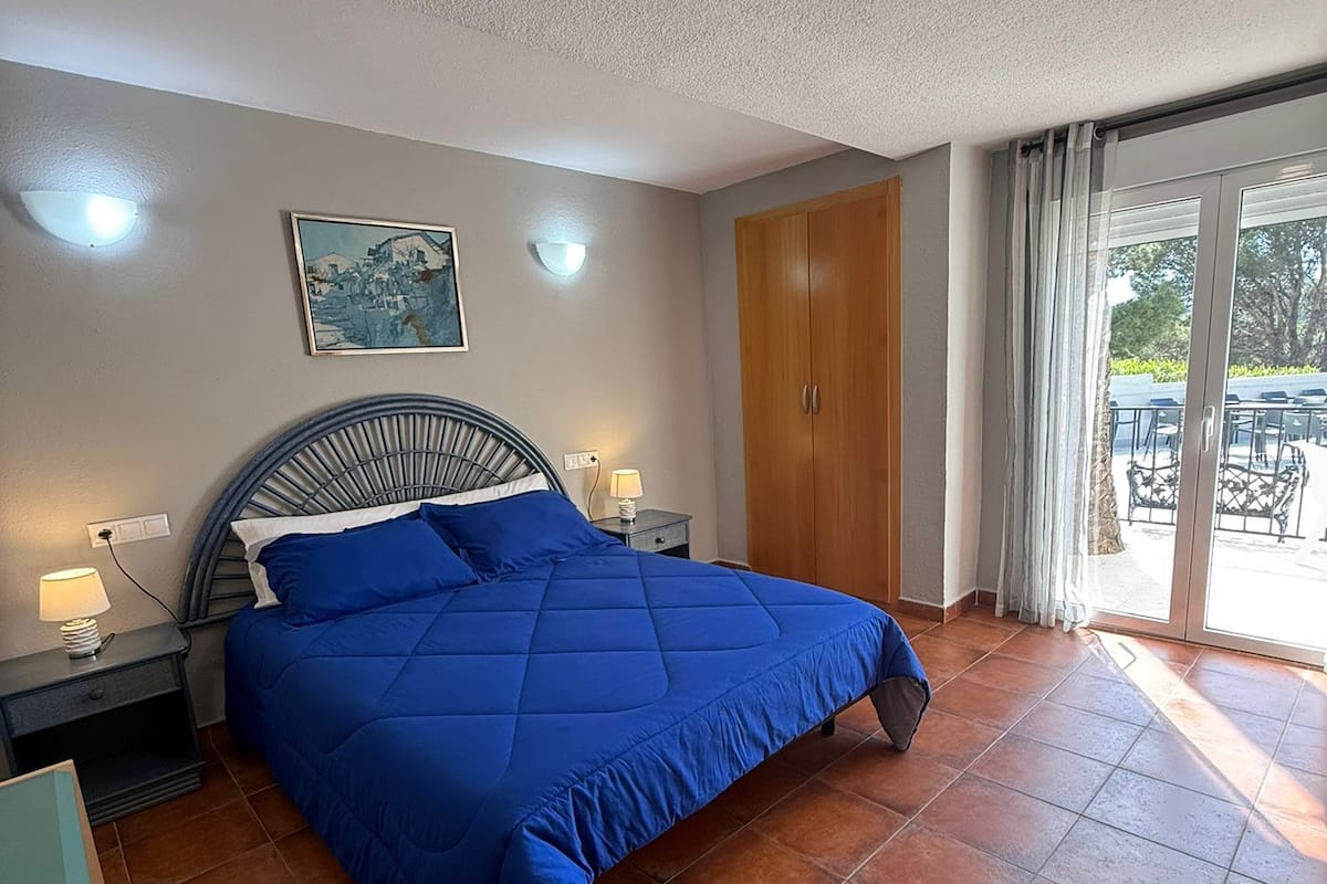 A bedroom with a large bed, a nightstand, a wall-mounted lamp, a ceiling light, a wooden wardrobe, and a sliding glass door leading to an outdoor area.