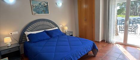 Un dormitorio con una cama grande, una mesita de noche, una lámpara de pared, una luz de techo, un armario de madera y una puerta de vidrio corredera que da a un espacio exterior.