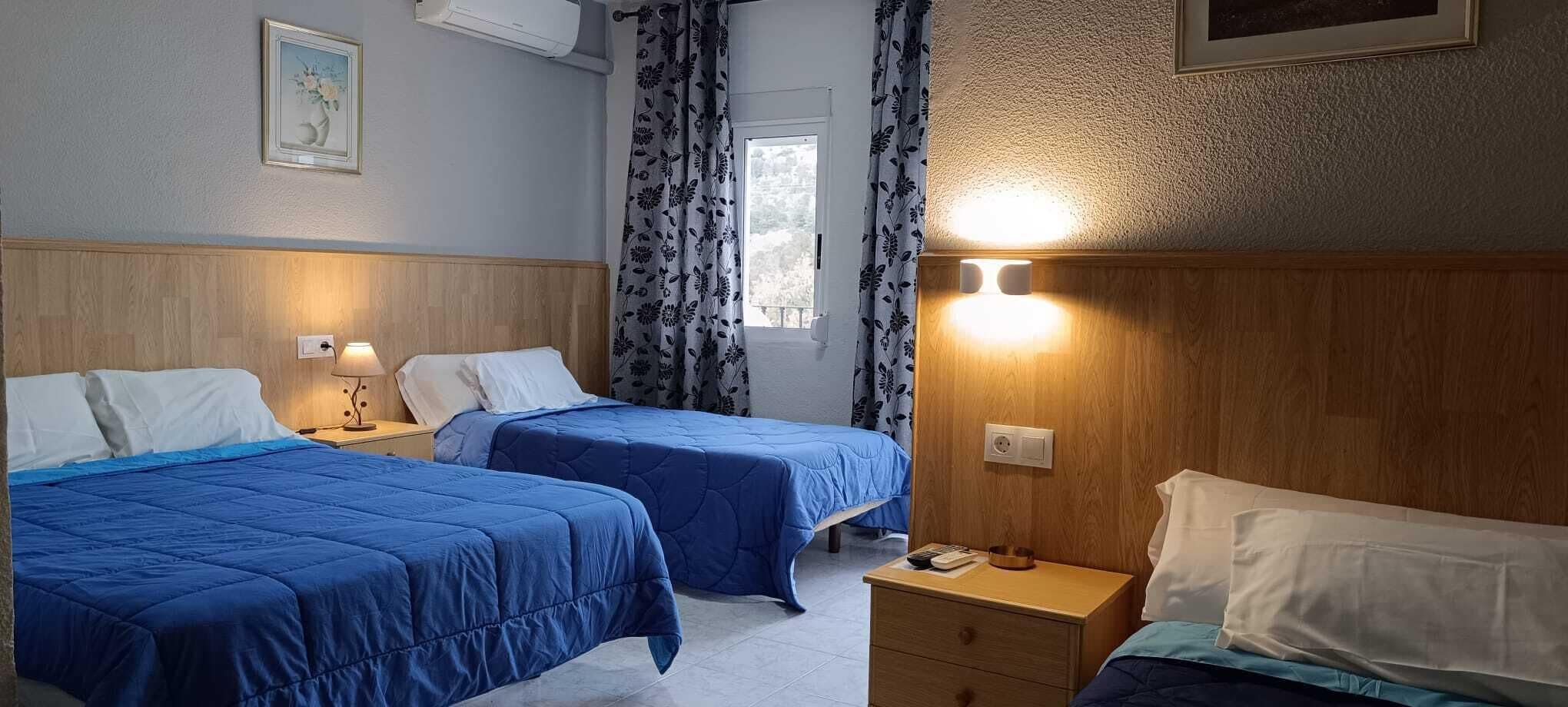 A hotel room with two beds, a bedside table with a lamp, a window with curtains, and a wall-mounted air conditioner.