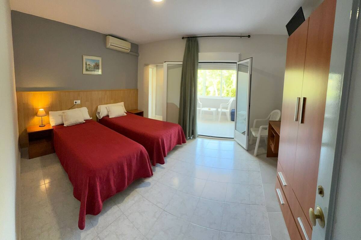 A hotel room with two beds, a nightstand, a wardrobe, and a balcony with chairs.
