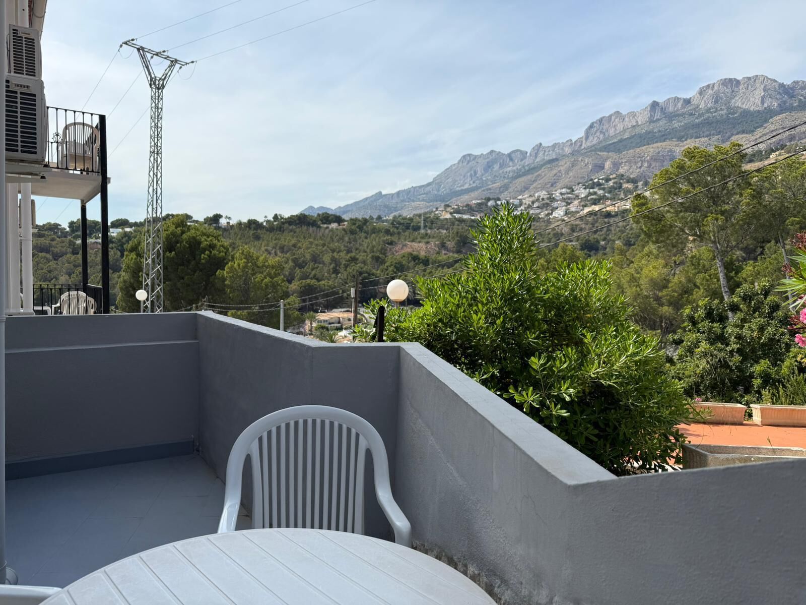 Uma varanda com cadeira branca, mesa e vista para montanhas e vegetação.