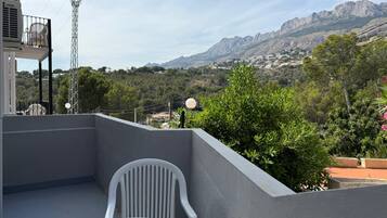 Uma varanda com cadeira branca, mesa e vista para montanhas e vegetação.