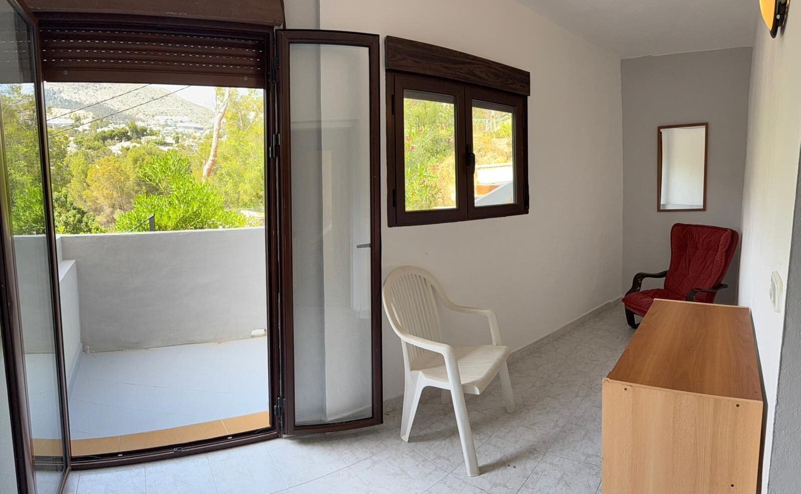 A room with a sliding glass door, a white plastic chair, a red chair, and a wooden table.