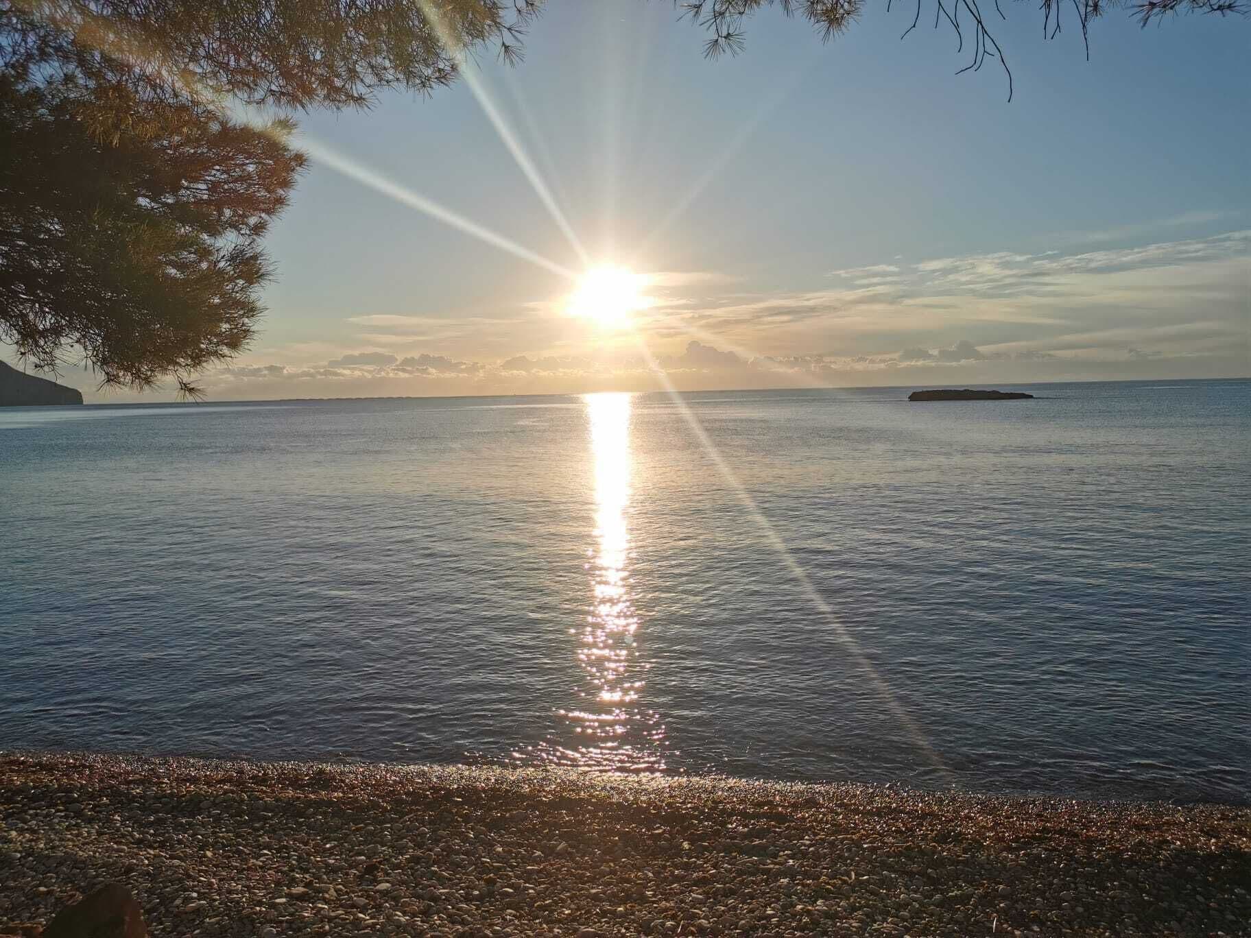 Une étendue d’eau calme, dont la surface reflète la lumière du soleil, bordée d’une côte rocheuse et entourée de reliefs lointains.