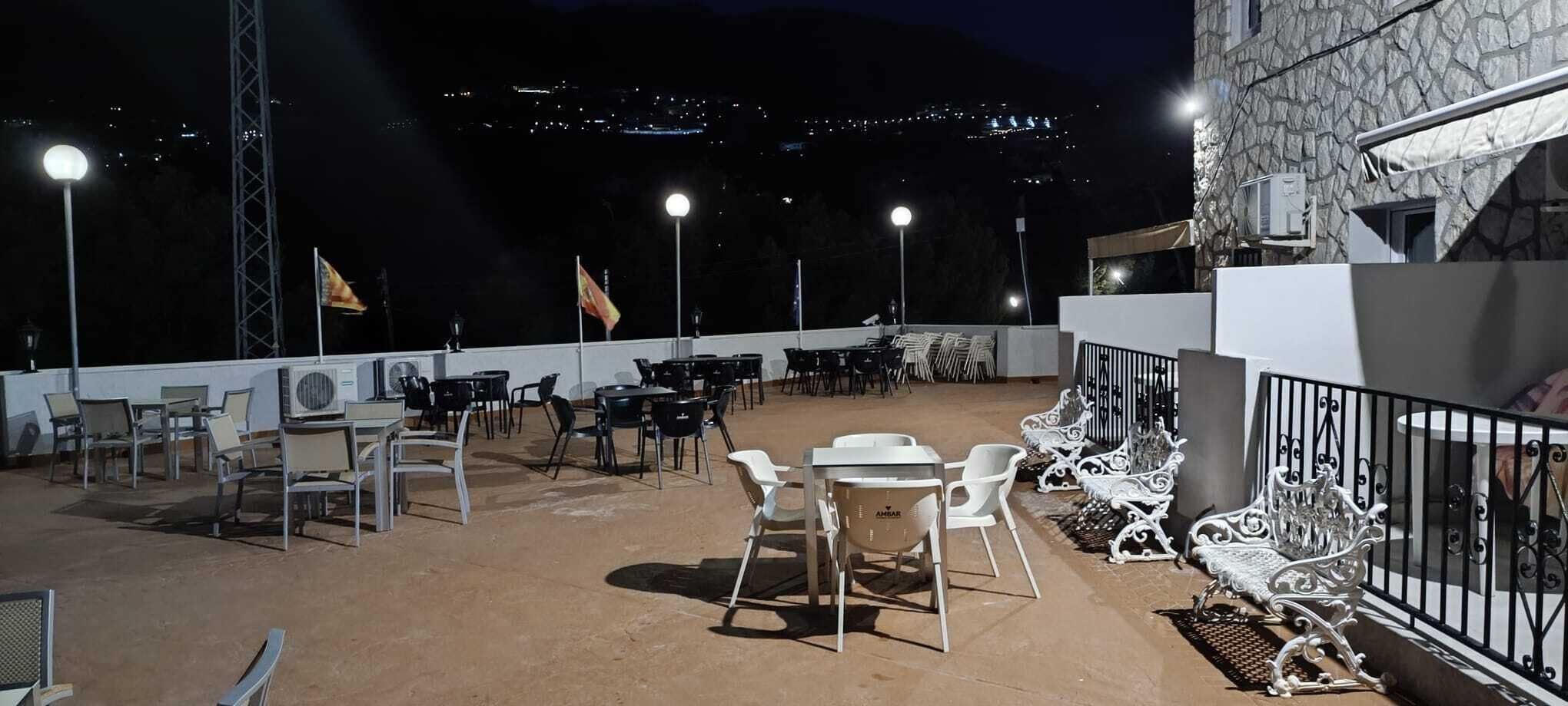An outdoor terrace with tables and chairs, a stone wall, and a view of the city at night.