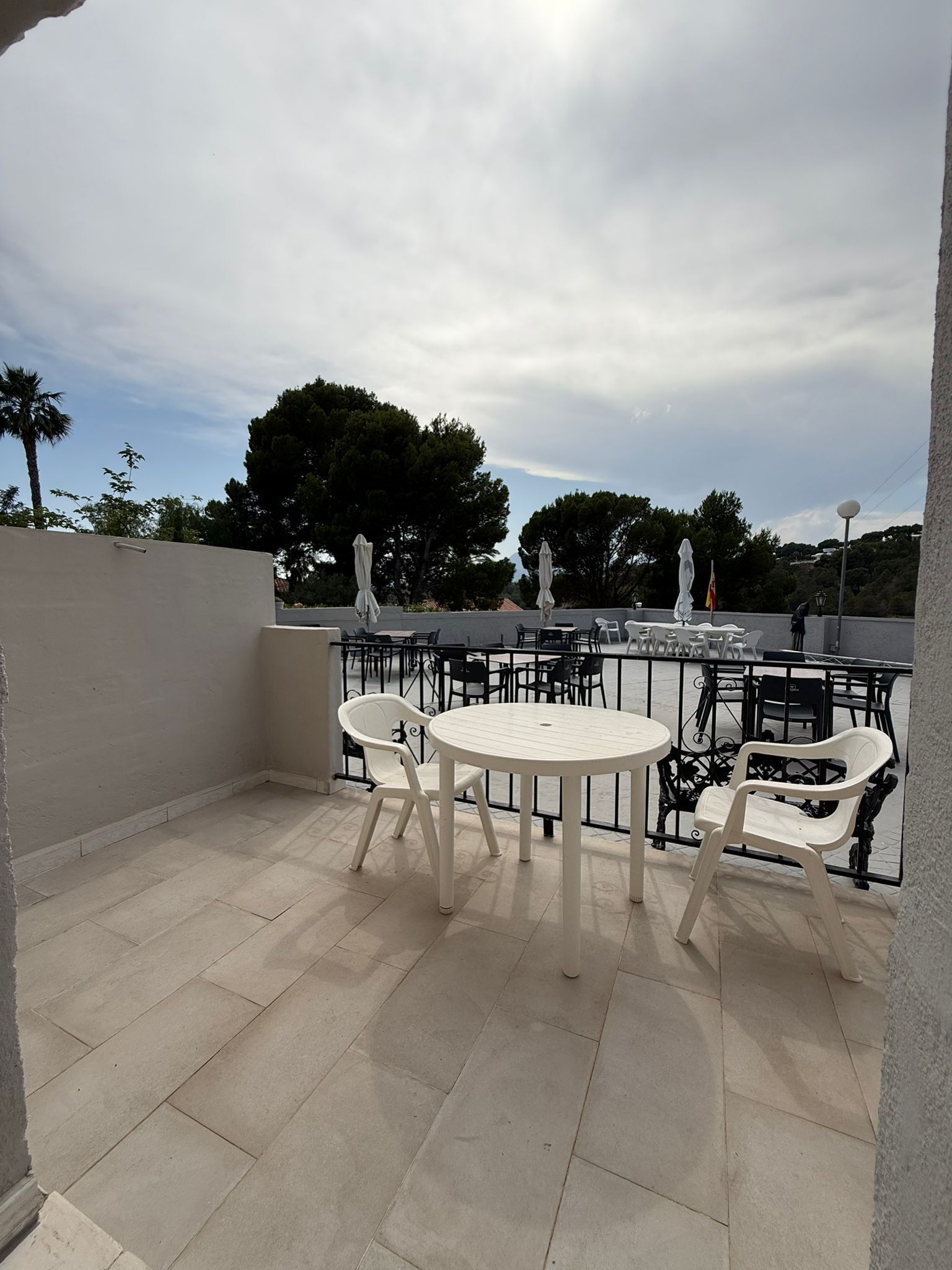 An outdoor patio with a round table and chairs, a railing, and a view of trees and buildings in the background.