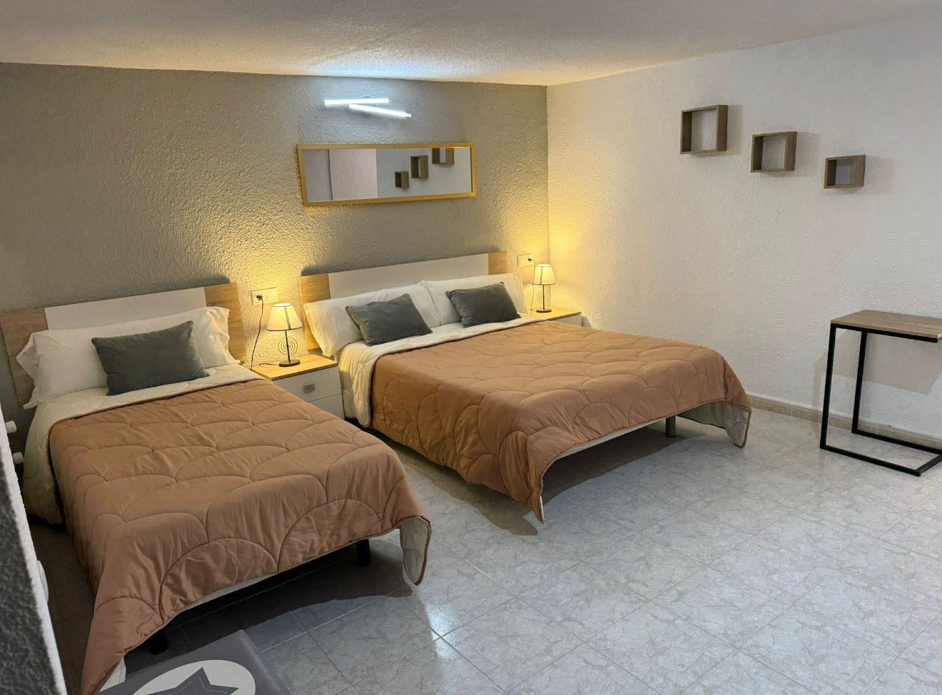 A hotel room with two beds, a bedside table with a lamp, a small table, and a mirror on the wall.