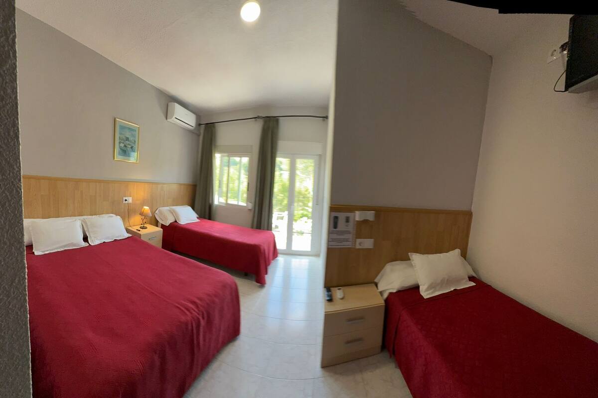 A hotel room with two single beds, a wooden headboard, a bedside table with a lamp, a window with curtains, and a view of greenery outside.