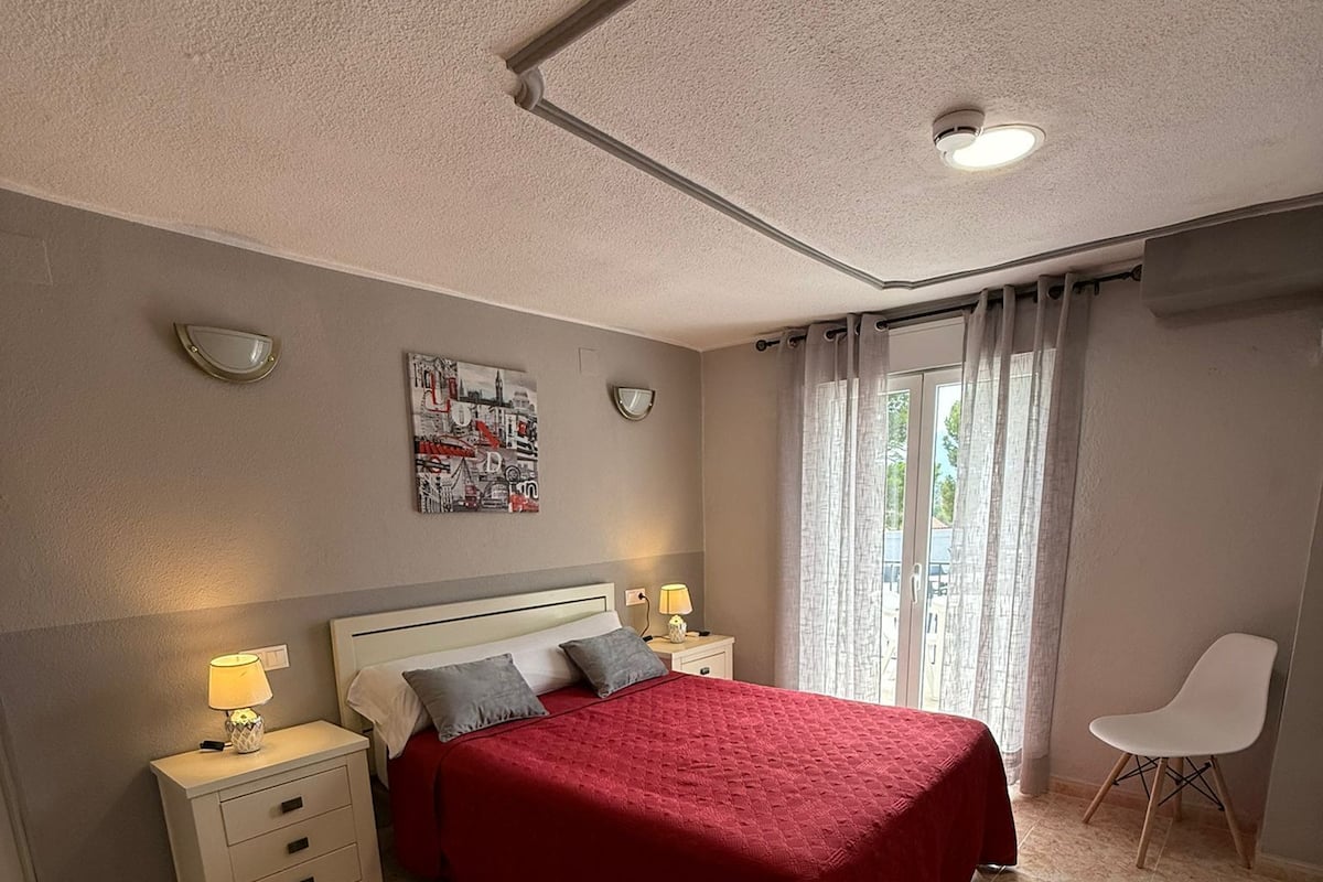 A bedroom with a bed, nightstands, a chair, a window with curtains, and a wall-mounted picture.