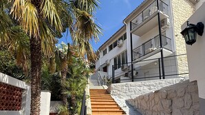 Una escalera que lleva a un edificio con balcón, rodeado de palmeras y un muro de piedra.