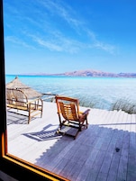 Una terraza de madera con dos sillas de madera mirando un mar tranquilo y colinas lejanas.