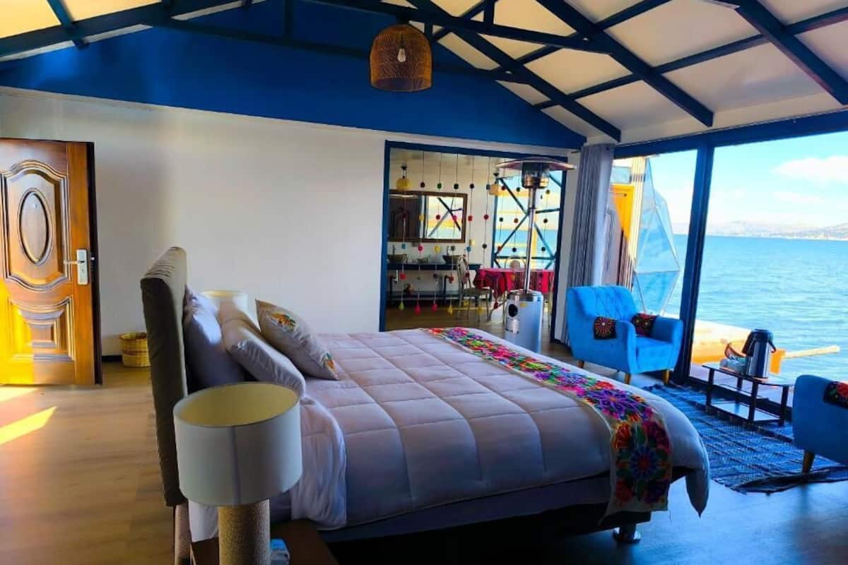 A bedroom with a bed, bedside lamps, a chair, and a view of the sea.