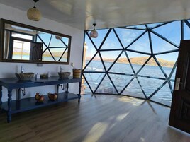 Una habitación con un ventanal grande, suelo de madera, una mesa azul con cuencos y un espejo.