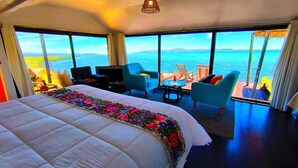 A hotel room with a large bed, a TV, a sofa, and a small table. There are large windows with a view of the sea. - uros killa lodge (Puno)