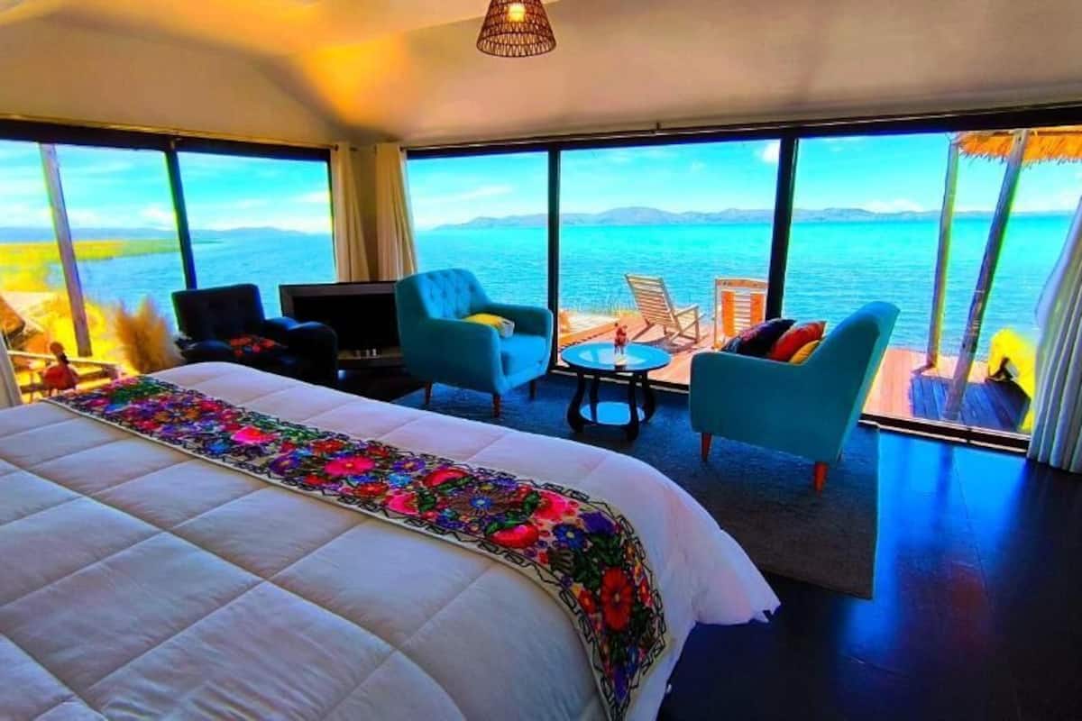 A hotel room with a large bed, a TV, a sofa, and a small table. There are large windows with a view of the sea.