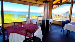 An open-air dining area with tables and chairs, a large window with a view of the water and hills, and a thatched roof structure. - uros killa lodge (Puno)