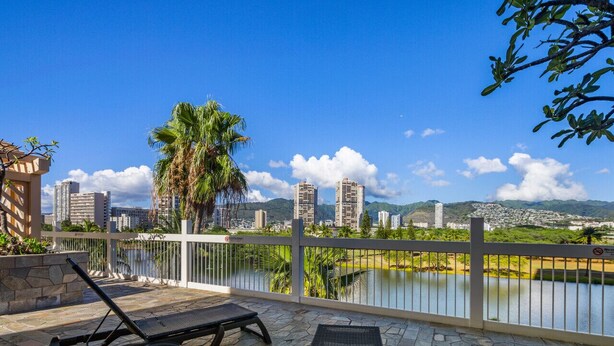 Uma varanda com duas cadeiras de descanso, piso de pedra, guarda-corrimão branco e vista para a paisagem urbana e um lago.