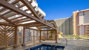Piscina com pergolado de madeira, posto de salva-vidas e área à beira da piscina com vista para edifícios próximos.