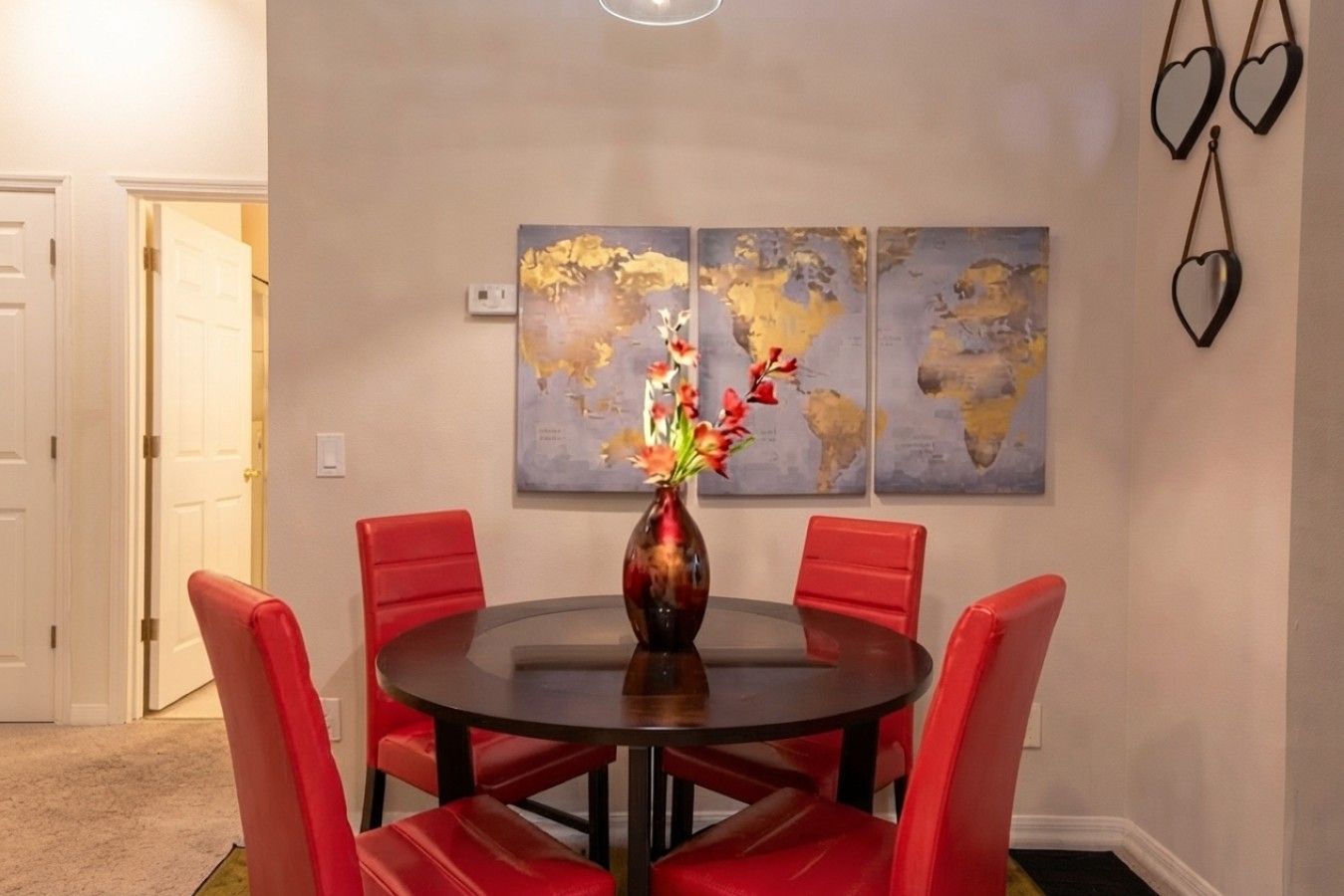 Een eethoek met een ronde tafel, rode stoelen, een vaas met bloemen en twee wereldkaarten aan de muur.