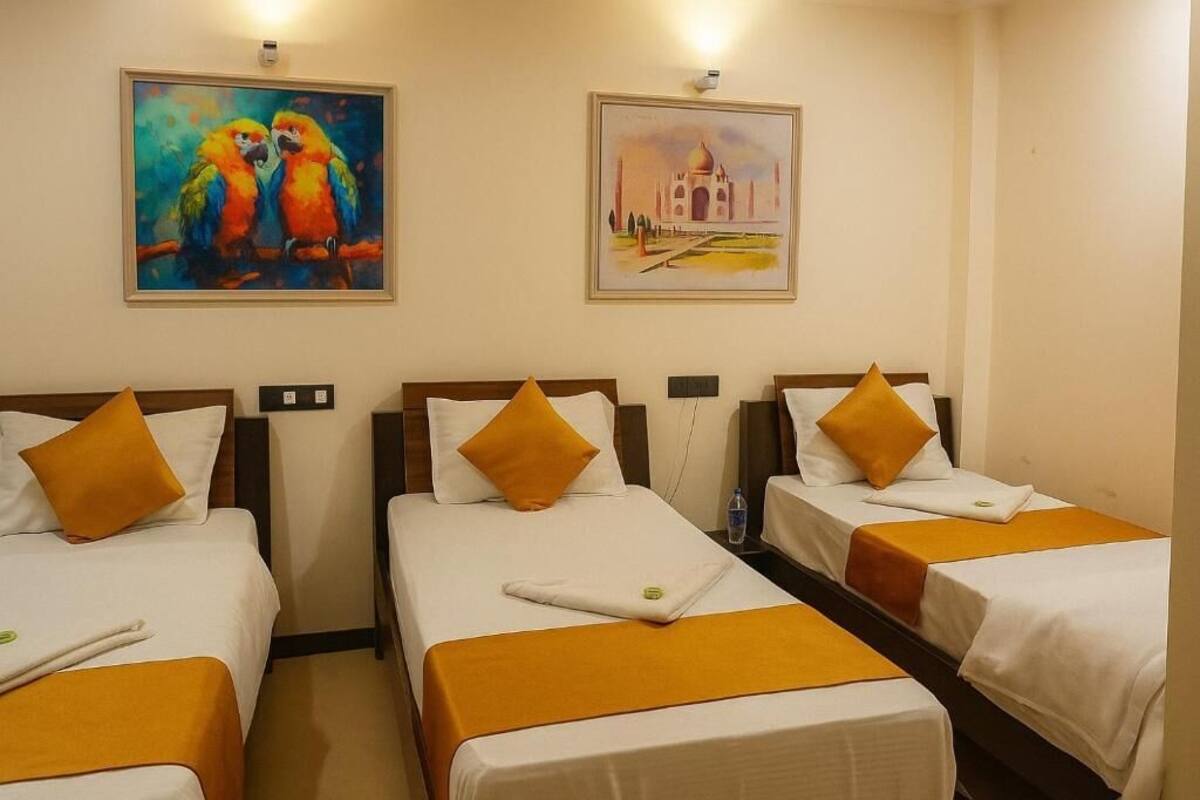 A hotel room with three single beds, each with a white bedspread and an orange throw blanket. There are two paintings on the wall, one of parrots and another of a building.
