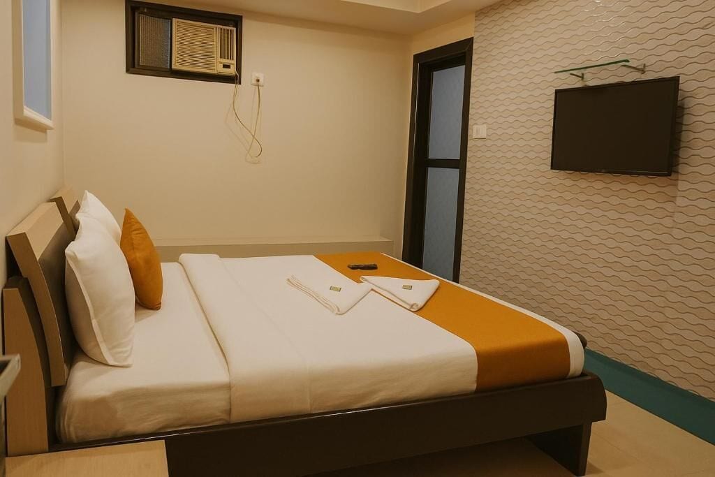 A hotel room with a bed, a television mounted on the wall, and a window with an air conditioning unit.