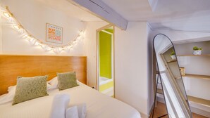 A hotel room with a bed, a headboard, a wall-mounted light fixture, a mirror, and a door leading to a bathroom. - L'Ô en Couleur - Au coeur de Bordeaux (Bordeaux)