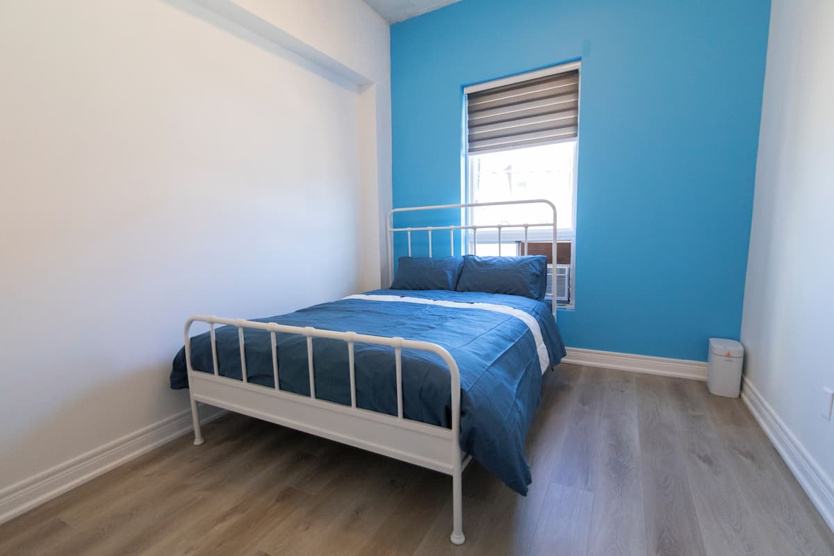 Une chambre à coucher avec un lit, une fenêtre avec des stores et une poubelle.