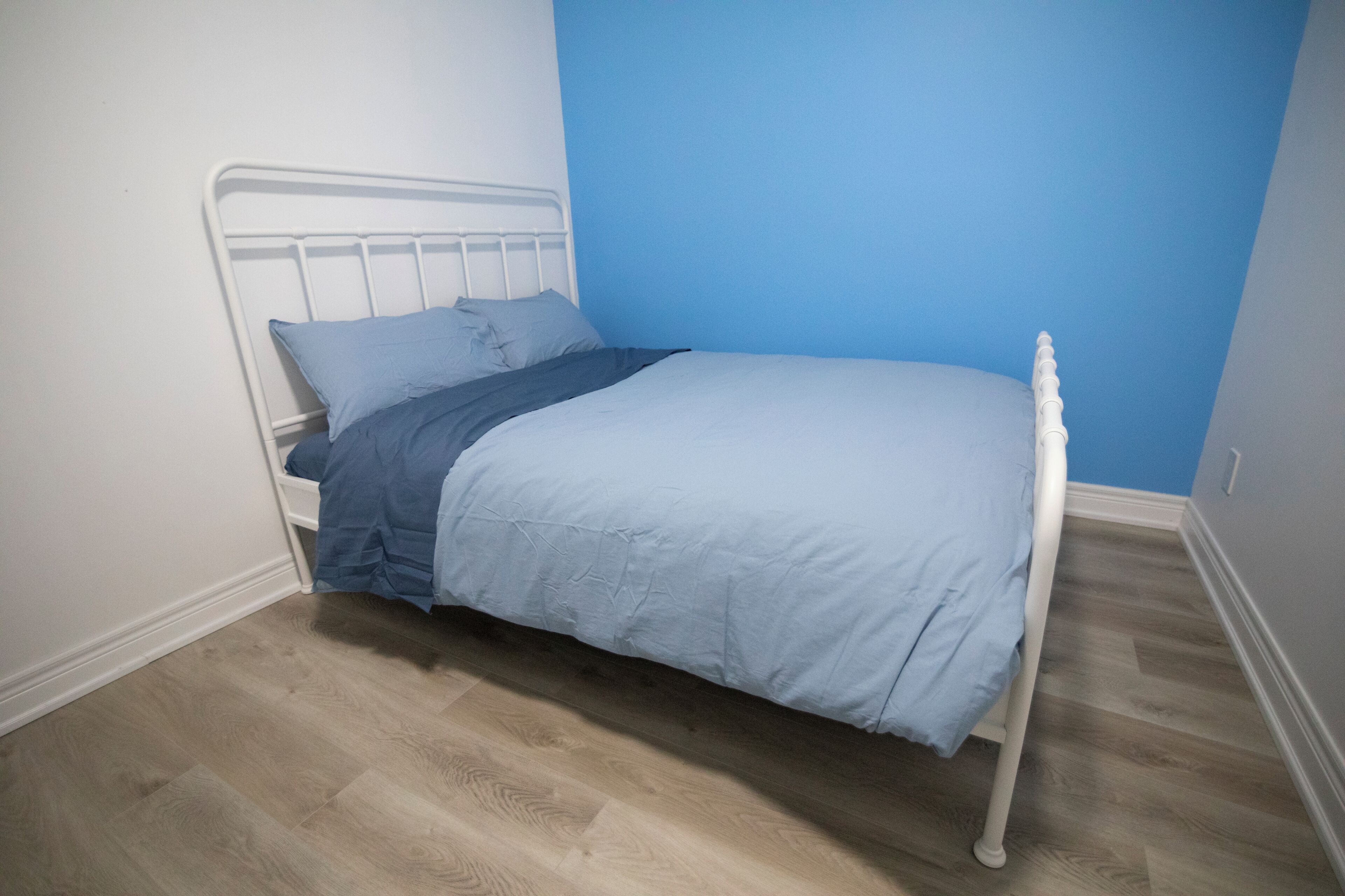 Una cama individual con estructura metálica blanca y colcha azul en una habitación con suelo de madera clara y una pared de acento azul.